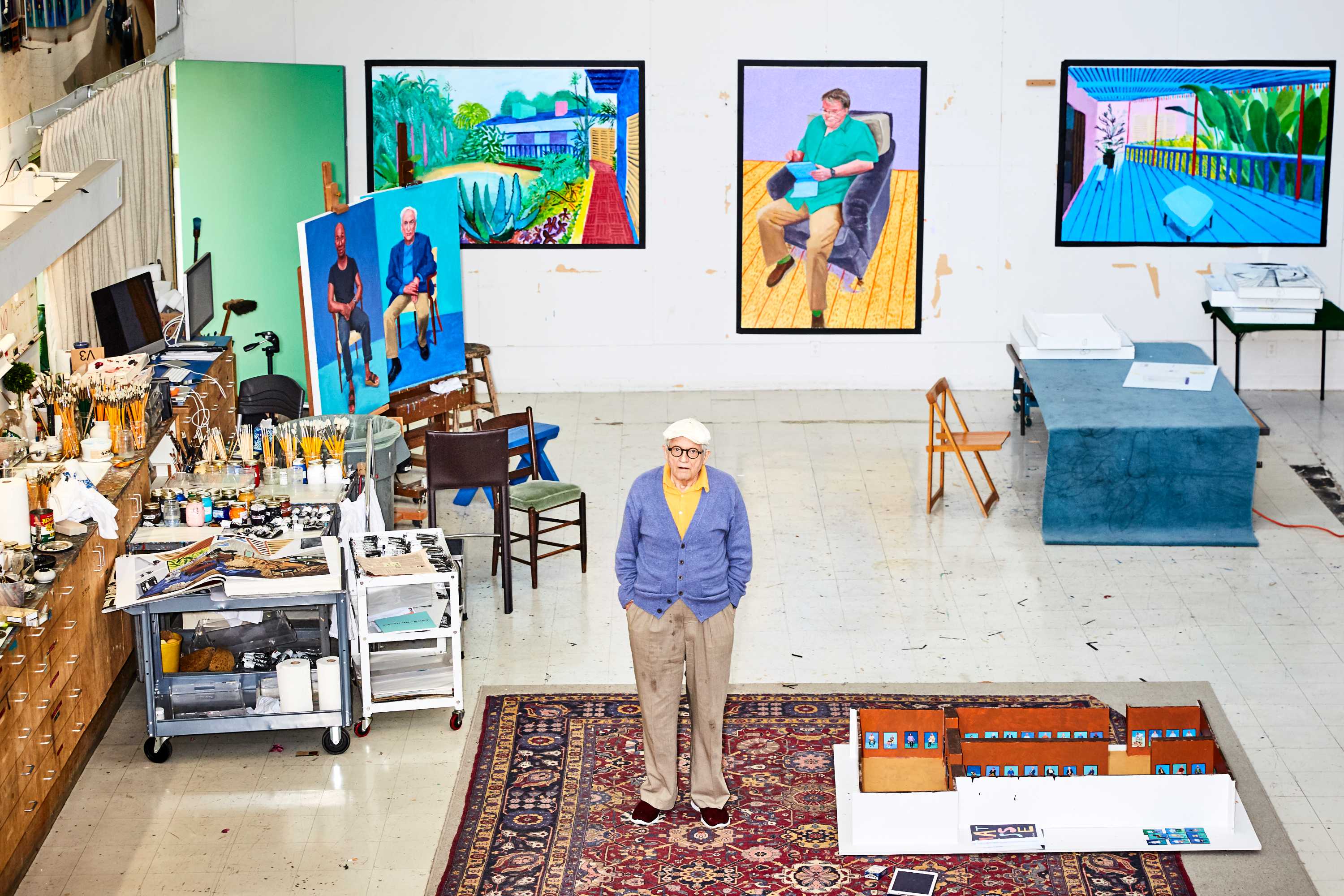 A shot from above of David Hockney standing in a warehouse type studio with paintings on the walls and lots of paints on a shelf