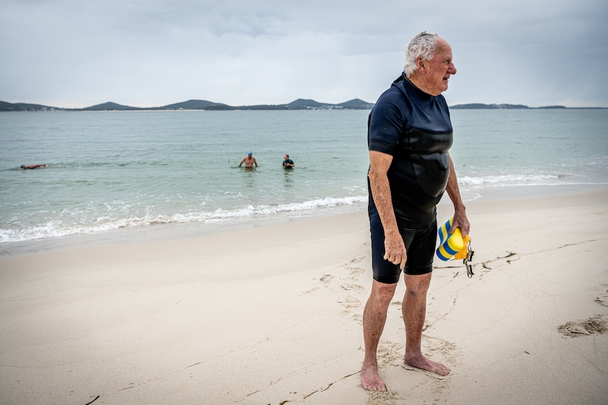 a older man in a wet suit stands at the ocean shore