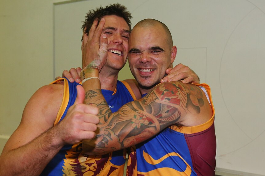 Two smiling teammates from the Brisbane Lions AFL side stand with their arms around each other in the rooms after a win.