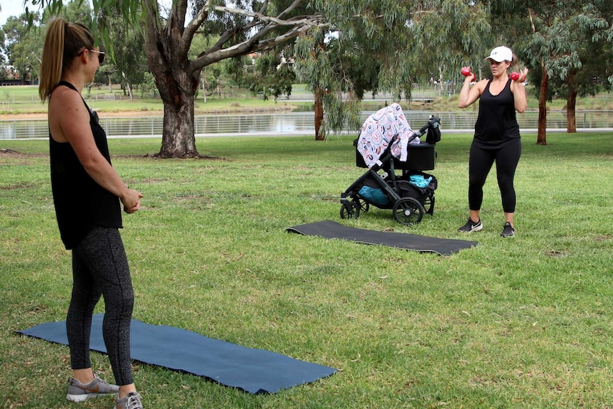 Two women in active wear stand three meters apart as they work out in a park. One woman stands next to a pram holding weights.