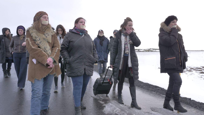 A group of women walk along a path in a cold, snowy environment with one woman pulling a suitcase behind her.