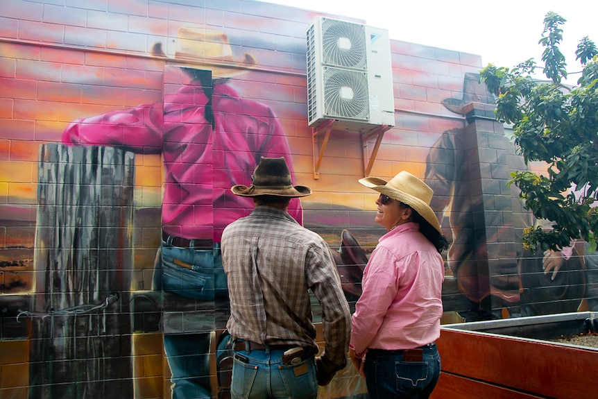 A man and woman stand in front of a mural depicting them sitting on a wooden fence.