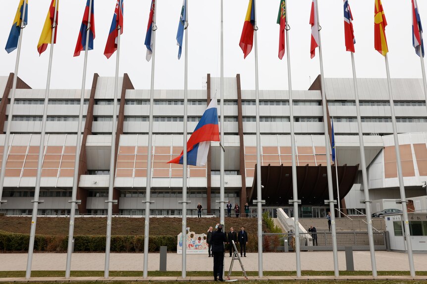 Thirteen flags are raised in a line in front of a building. Russia's, in the middle, is being lowered by an official in a suit
