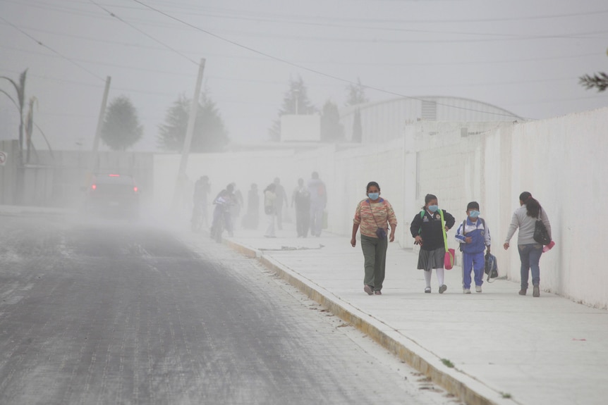 Four people wearing masks walking along a street covered with ash and ash visible in the air. 