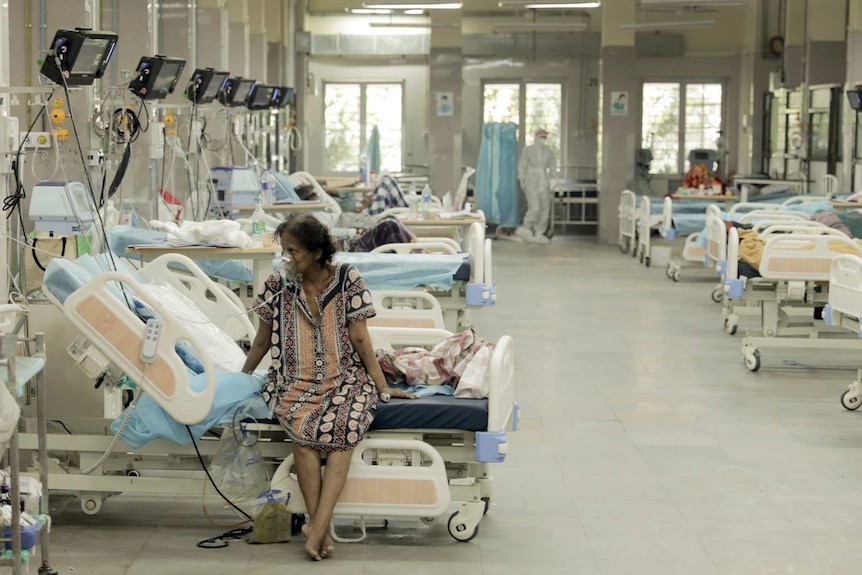 A woman wearing a dress sits on a bed with a cup of her faces giving her air as she is surrounded by hospital beds