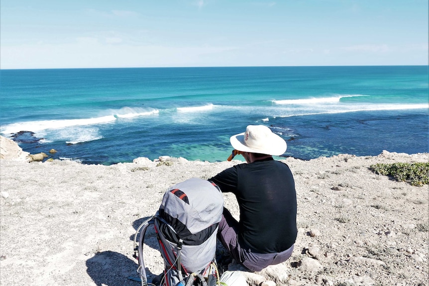 Man sitting on cliff with back to camera, back pack on ground looking at blue ocean waves