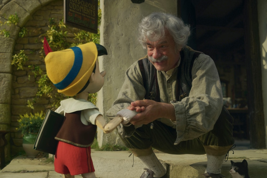 Grey haired older man kneeling and interacting with small humanoid toy wearing yellow hat, red shorts, white and brown top. 