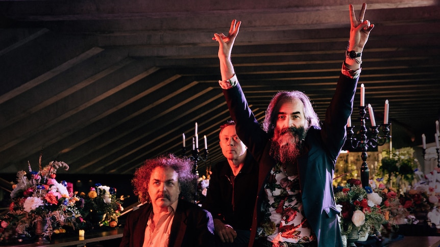 three members of band dirty three in jackets sit in a dark backstage area. warren ellis has arms aloft and holds peace signs