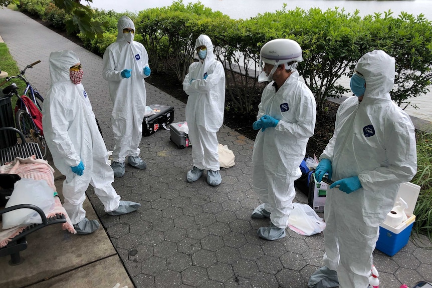Five people wearing white protective suits, glooves, masks, goggles and feet coverings standing on street near a river.