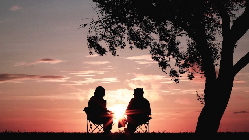 A couple sitting on camp chairs watch a pink sunset.