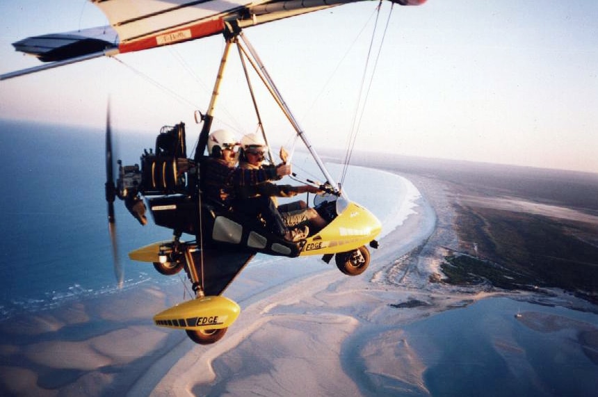 Two people fly in an ultra-light aircraft over a beach at sunset.