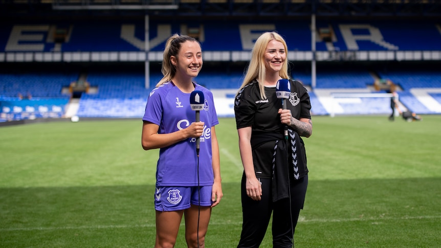 A woman soccer player wearing blue holds a microphone next to an interviewer in a stadium
