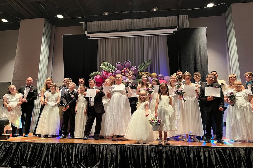 Group of debutantes in white dresses stand with partners in suits on stage, smiling at cameras.