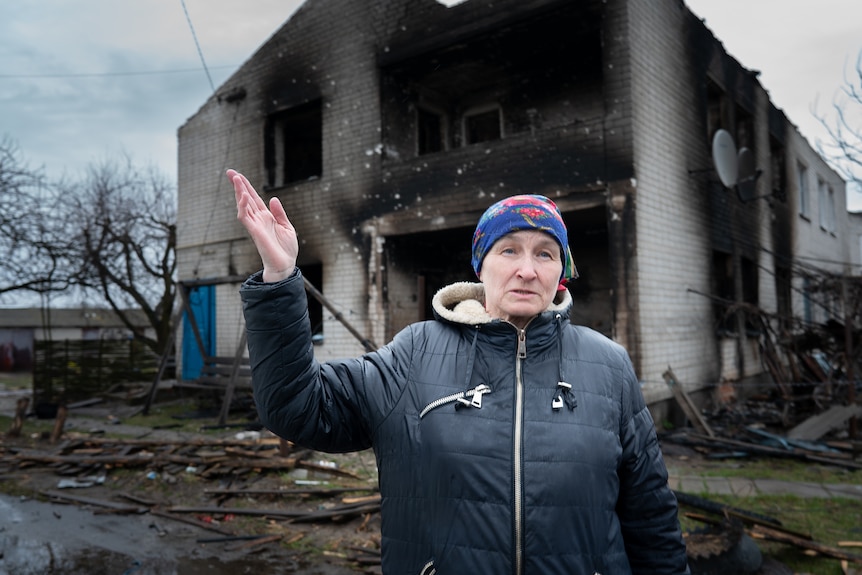A woman in a blue scarf and black puffer gestures outside a destroyed building. 