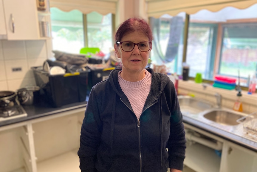 An older, bespectacled woman stands in a kitchen.
