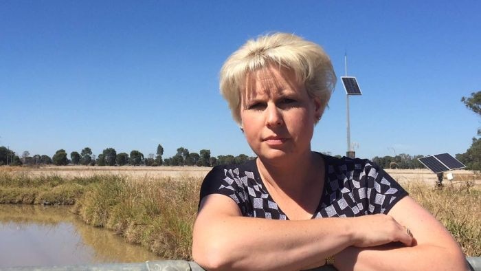 A woman with short blond hair leaning against a fence, with an irrigation channel in the background.