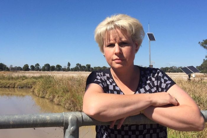 A woman with short blond hair leaning against a fence, with an irrigation channel in the background.
