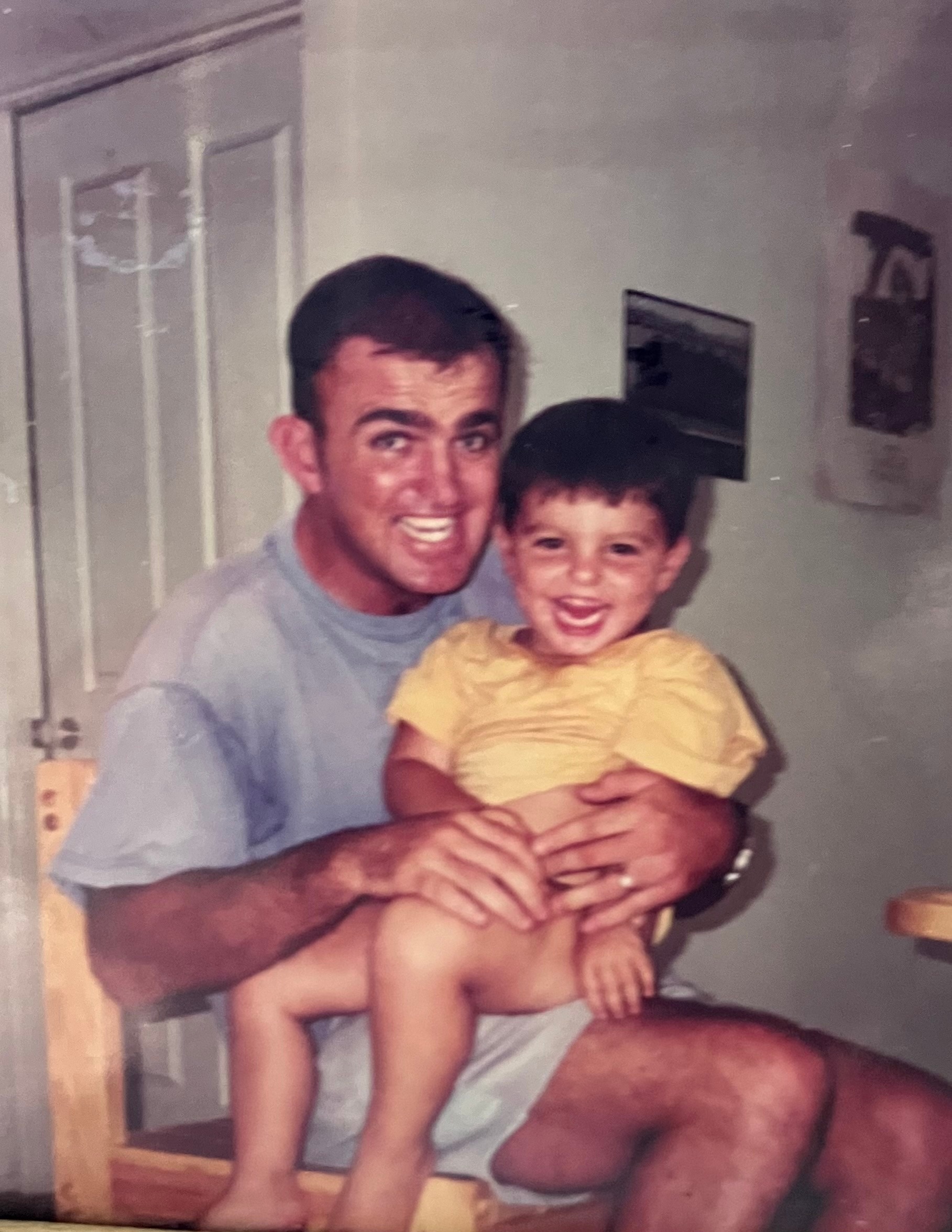 A photo of a smiling man holding a little boy in a yellow shirt.