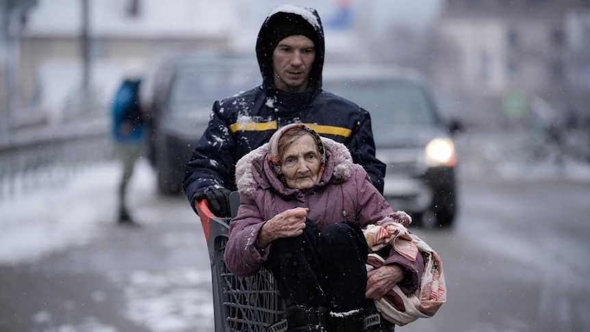 An elderly woman wearing a purple jumper is carried by a young man walking down a street full of snow