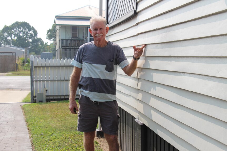 A man with white hair and a moustache points to a weatherboard on the side of a house