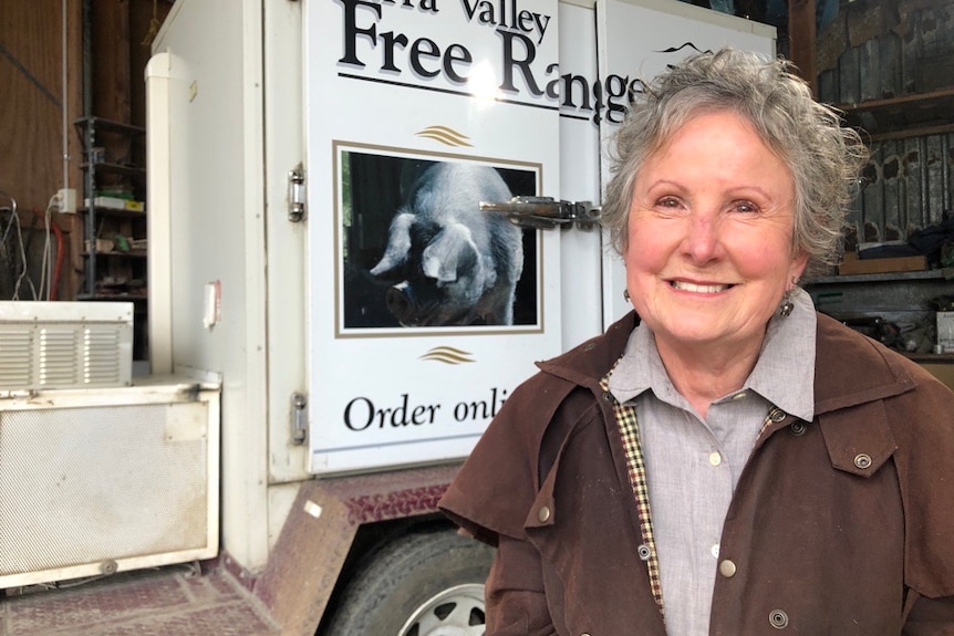 An older woman with short grey hair, wearing a brown trench coat, stands in front of a van.