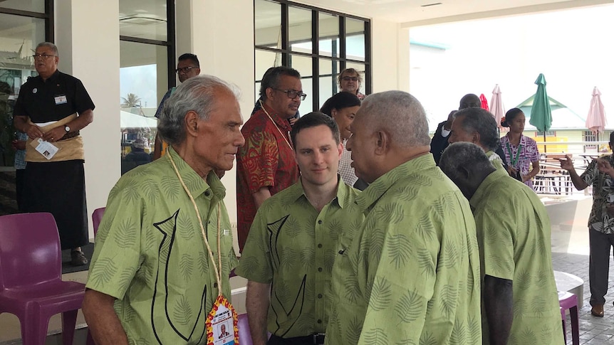 A group men wearing green shirts speak at the front of a building.