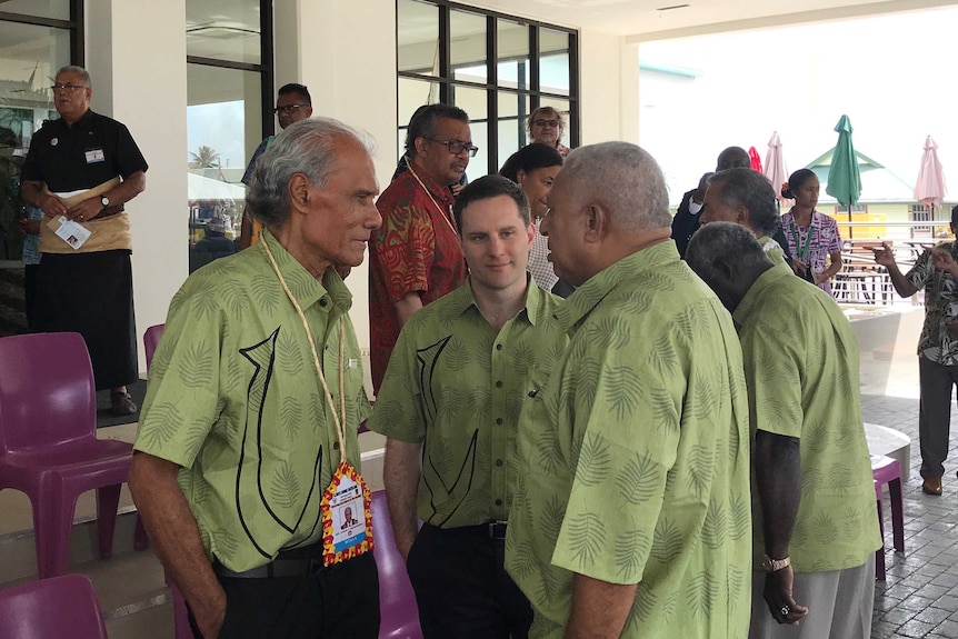 A group men wearing green shirts speak at the front of a building.