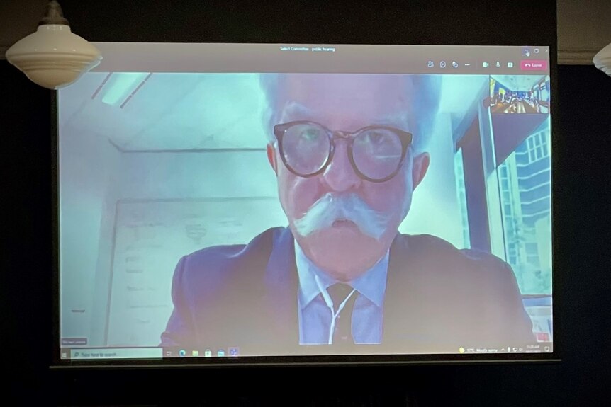 A man with glasses and a white moustache on a screen