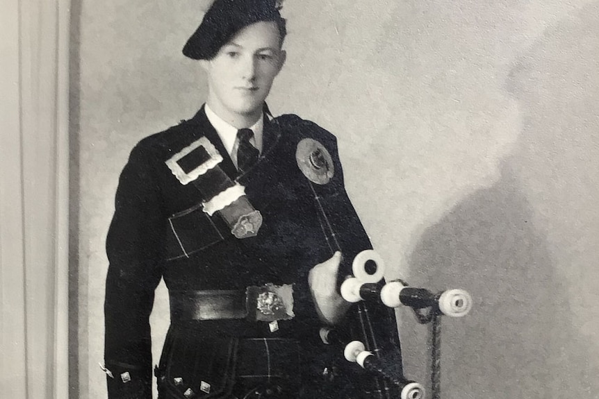 A young man stands formally for the camera in full pipe band costume — kilt, beret, sporran and pipes.