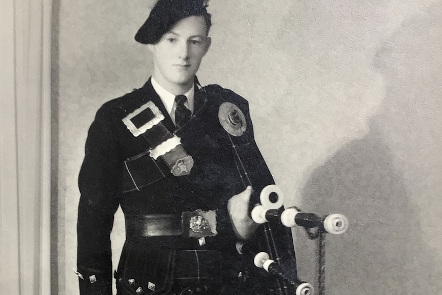 A young man stands formally for the camera in full pipe band costume - kilt, beret, sporran and pipes