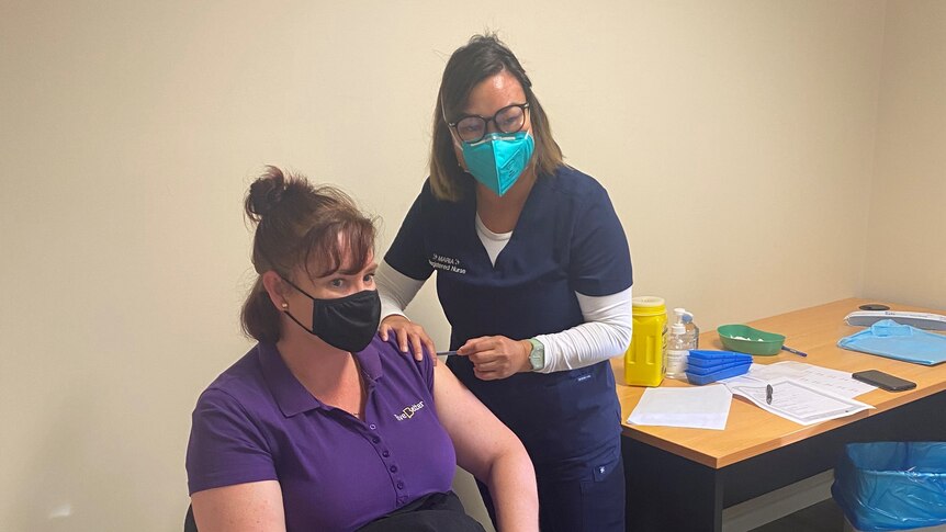 Two women with masks, one wearing purple receiving a vaccination from the other