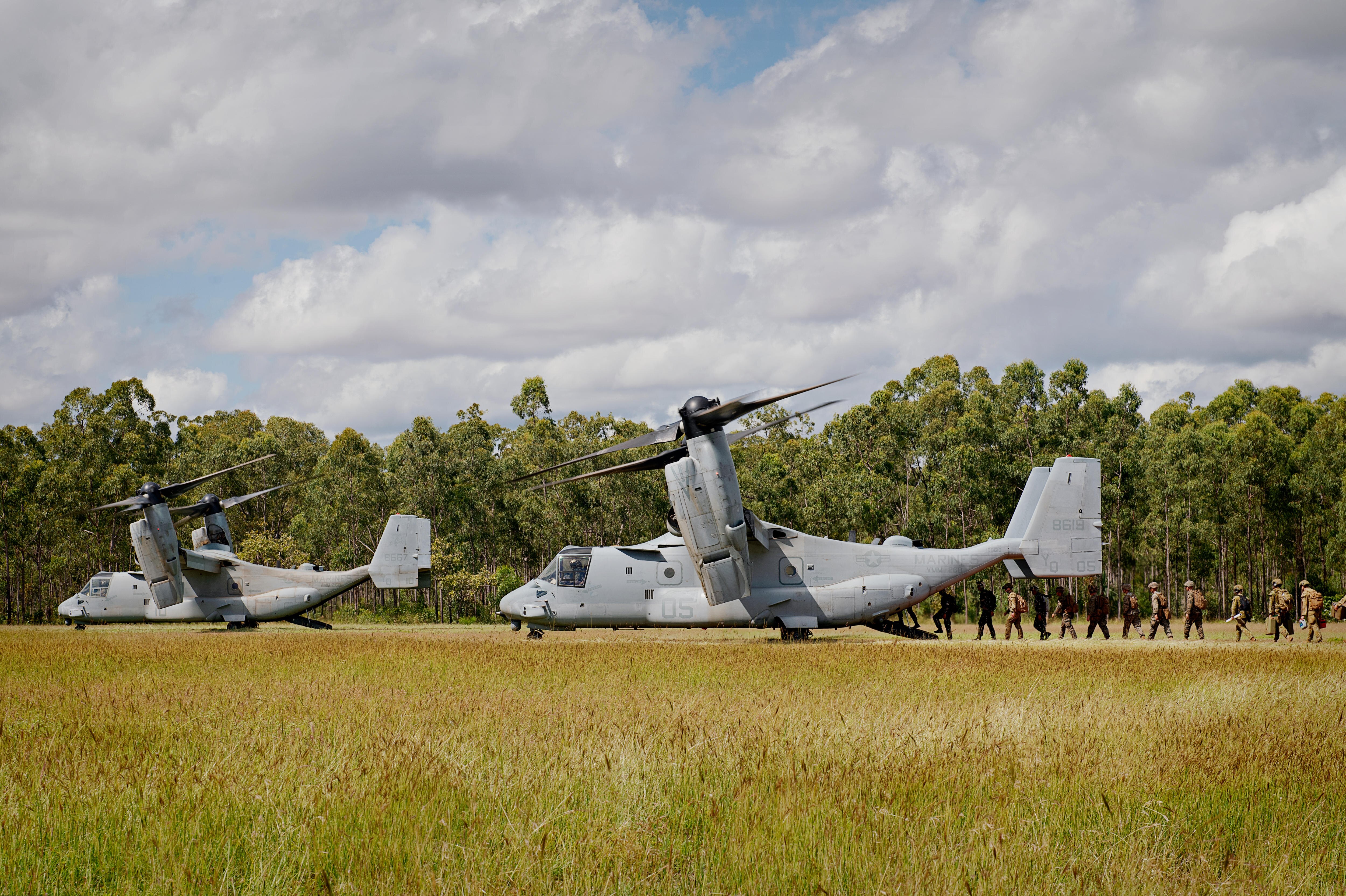 Two large military aircraft, in a grassy field, with a line of marines moving into the hatch of one