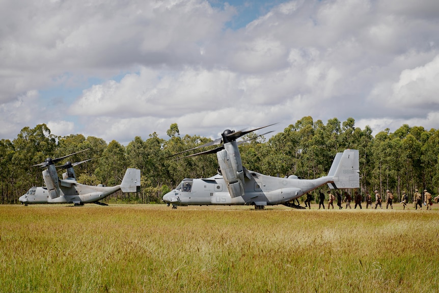 Two large military aircraft, in a grassy field, with a line of marines moving into the hatch of one