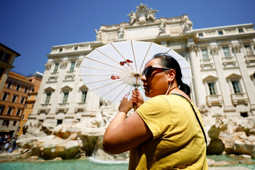 A woman wearing a yellow shirt holds up an umbrella to protect her from the sun as she stands next to a fountain.