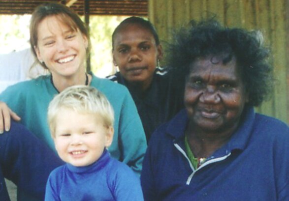 A woman wearing a blue jumper sits with her grandchildren beside her. They are all happy and smiling.