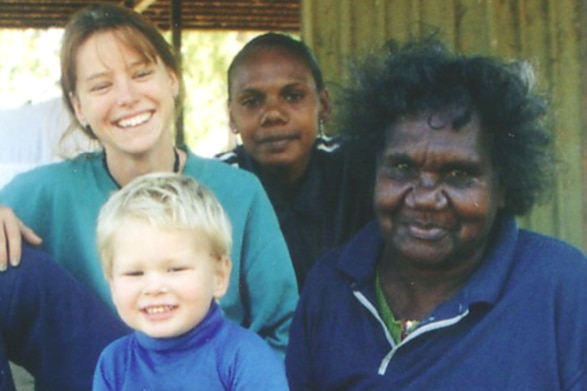 A woman wearing a blue jumper sits with her grandchildren beside her. They are all happy and smiling.