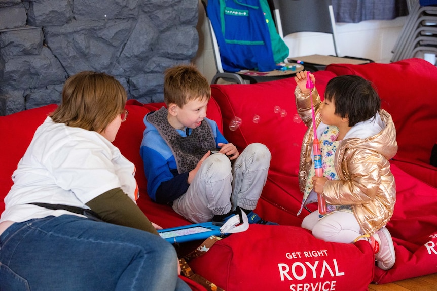 Three people sit on beanbags, two are children and one blows bubbles to another.