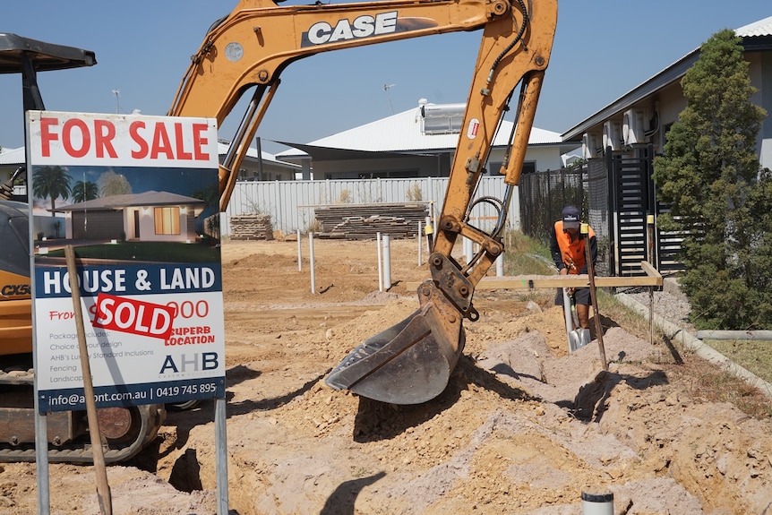 A digger is working on a block of land behind a 'For Sale' sign. The sign has a 'Sold' sticker on it.
