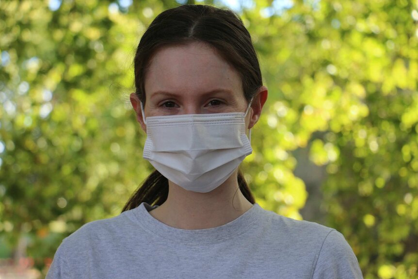 A dark haired woman with a mask looks at the camera while standing in a park.
