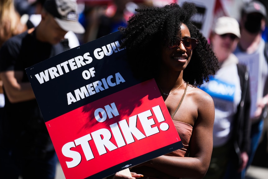 A Black woman holds a sign with Writers Guild of America on Strike written on it as she stands in a crowd.