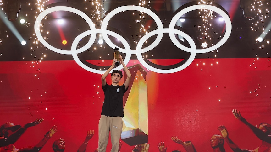 a teenag video gamer holding up a trophy over his head on stage in front of the olympic rings
