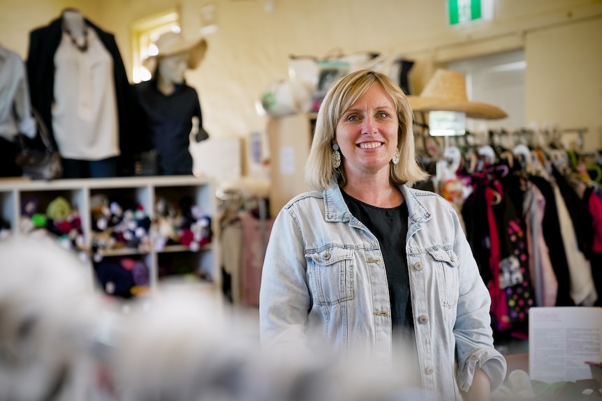 A blonde woman smiling in a shop full of clothes.