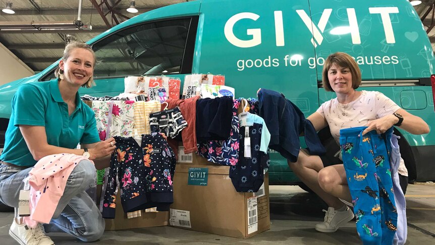 Two women hold up children's clothing in front of a green van with 'GIVIT goods for good causes' on it
