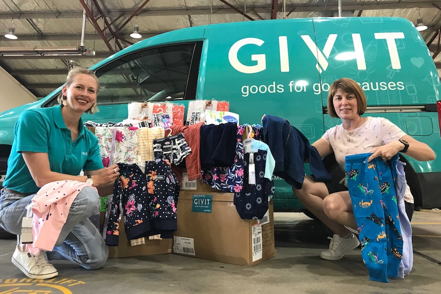 Two women hold up children's clothing in front of a green van with 'GIVIT goods for good causes' on it