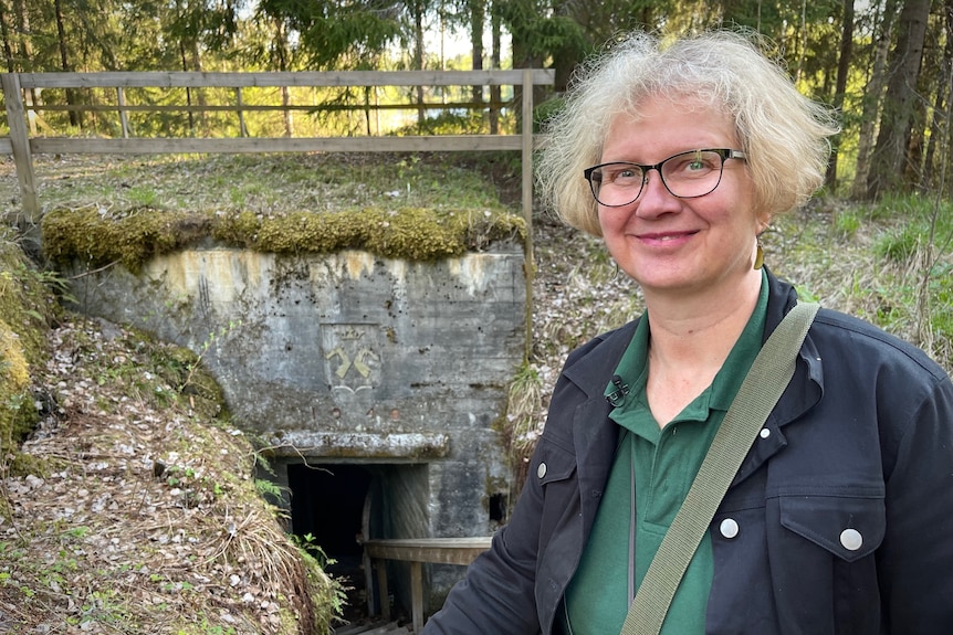 A blonde woman in glasses stands near the entrance to an old bunker.