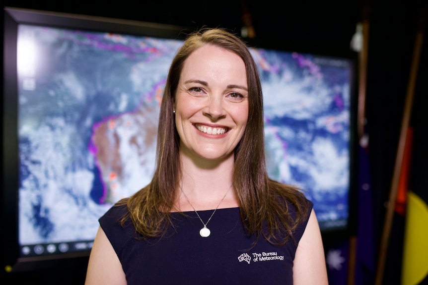 A woman smiling at the camera with a screen behind her showing clouds over Australia.