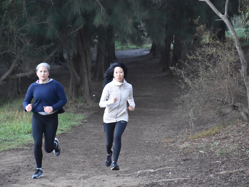 Two women jogging on a dirt track with trees behind them.