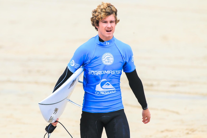 Man walking on beach holding a surfboard and wearing a blue competition top.