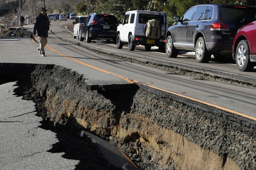 A large crack in a damaged road with a line of cars on top