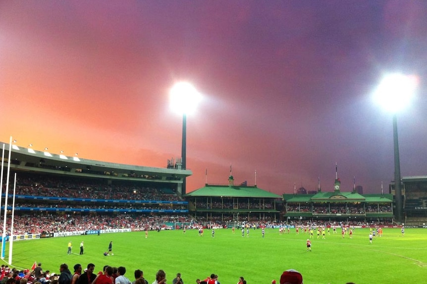 The setting sun shines through dark clouds sitting over the SCG during an AFL match.